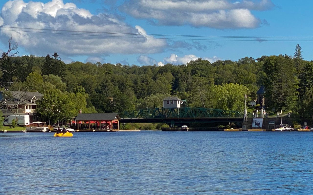 a view of the Muskoka river surrounded by tree with a cottage, dock, and bridge in the distance