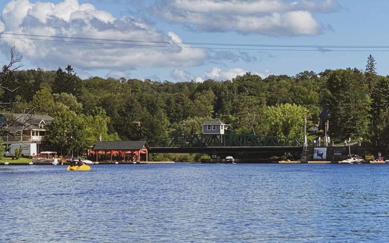 view of a Huntsville lake surrounded by trees with a cottage, dock, and bridge in the distance