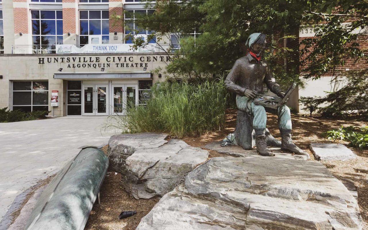 Tom Thompson bronze statue of a man sitting on a log with a paint palette in his lap in front of Huntsville Civic Centre, Algonquin Theatre