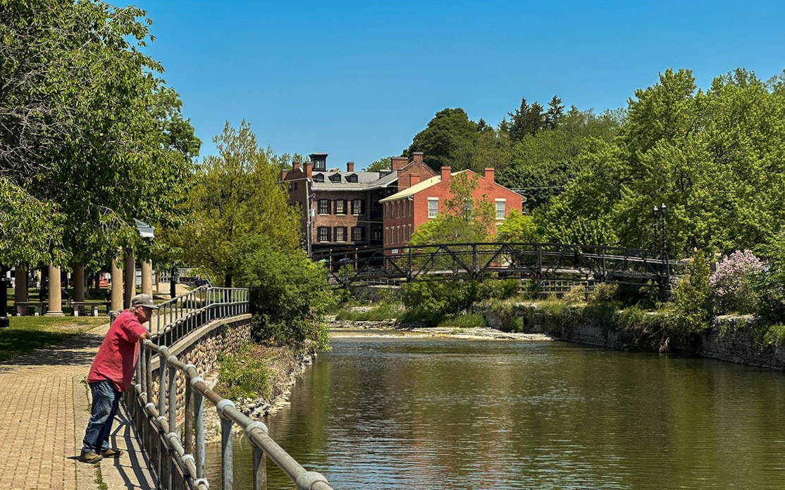 a man in a red shirt, jeans, and a ball cap overlooking a river from a walkway in Port Hope, Ontario